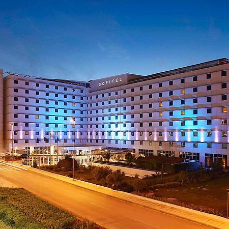 Sofitel Athens Airport Hotel Spata  Luaran gambar The photo shows a large modern hotel building, likely named "Sofitel," illuminated with blue and purple lights as evening approaches. The structure features multiple floors and a contemporary design, with large windows and a flat facade. In front of 