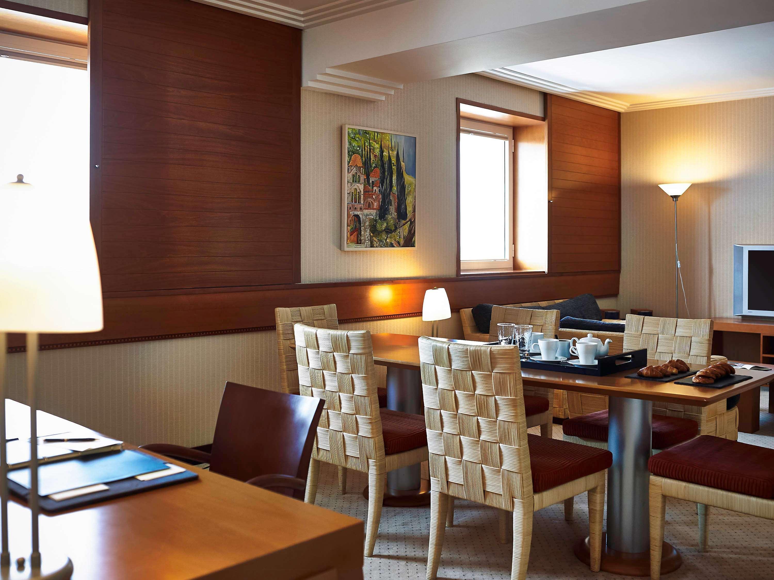 Sofitel Athens Airport Hotel Spata  Luaran gambar The photo shows a cozy dining area, likely in a hotel or a modern apartment. There is a wooden table surrounded by four chairs with woven backs. On the table, there's a tray with breakfast items, including some pastries. The walls are adorned with wa