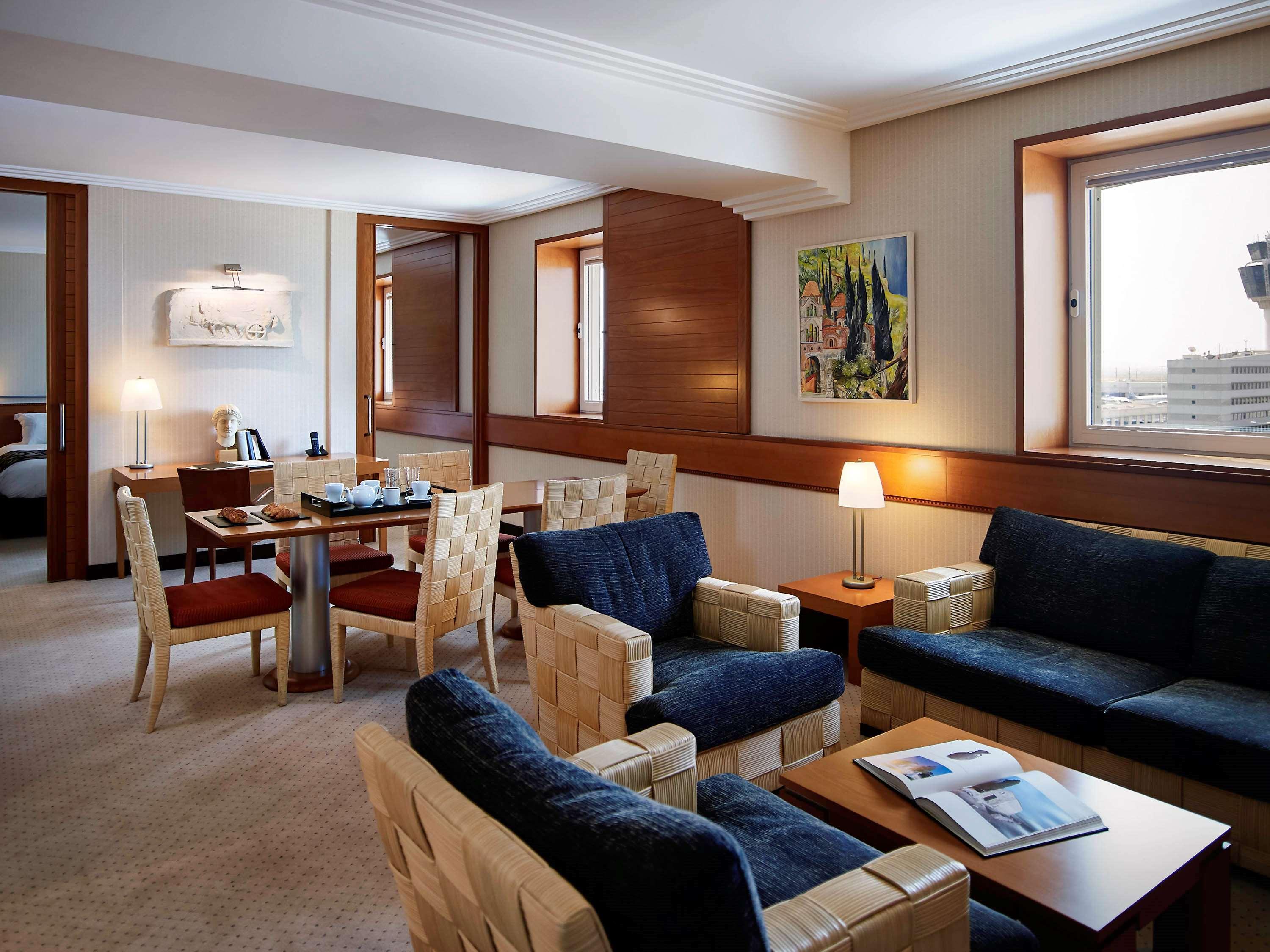 Sofitel Athens Airport Hotel Spata  Luaran gambar The photo shows a modern living area of a hotel suite. The room features a comfortable seating arrangement with dark blue sofas and armchairs, along with a wooden coffee table. There is a dining area with a table and chairs adjacent to the seating ar