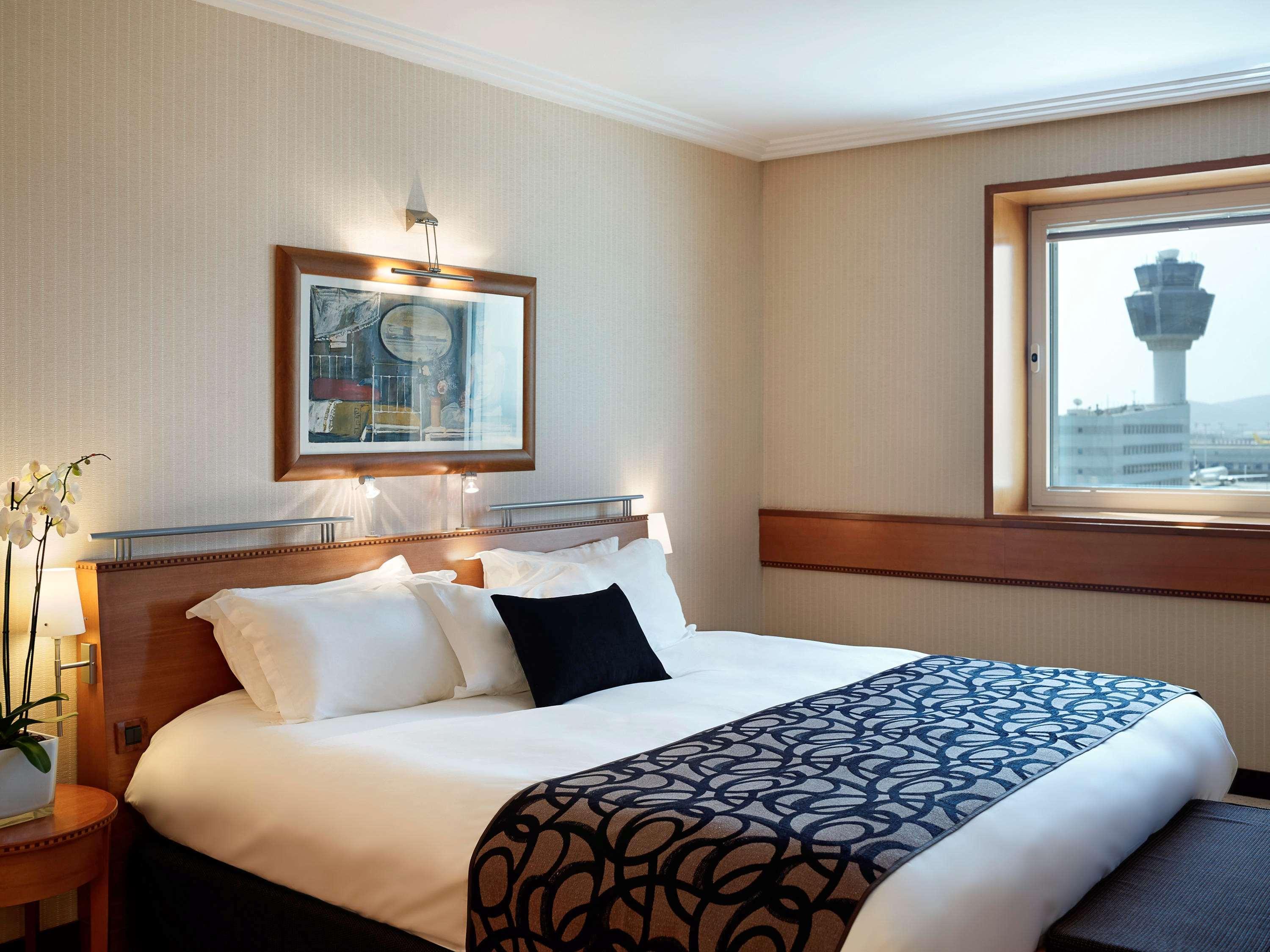 Sofitel Athens Airport Hotel Spata  Luaran gambar The photo shows a cozy hotel room with a large bed positioned against the wall. The bed is dressed in white linens with a decorative black and patterned throw. There are two pillows on the bed, along with one black accent pillow. 

The room features 