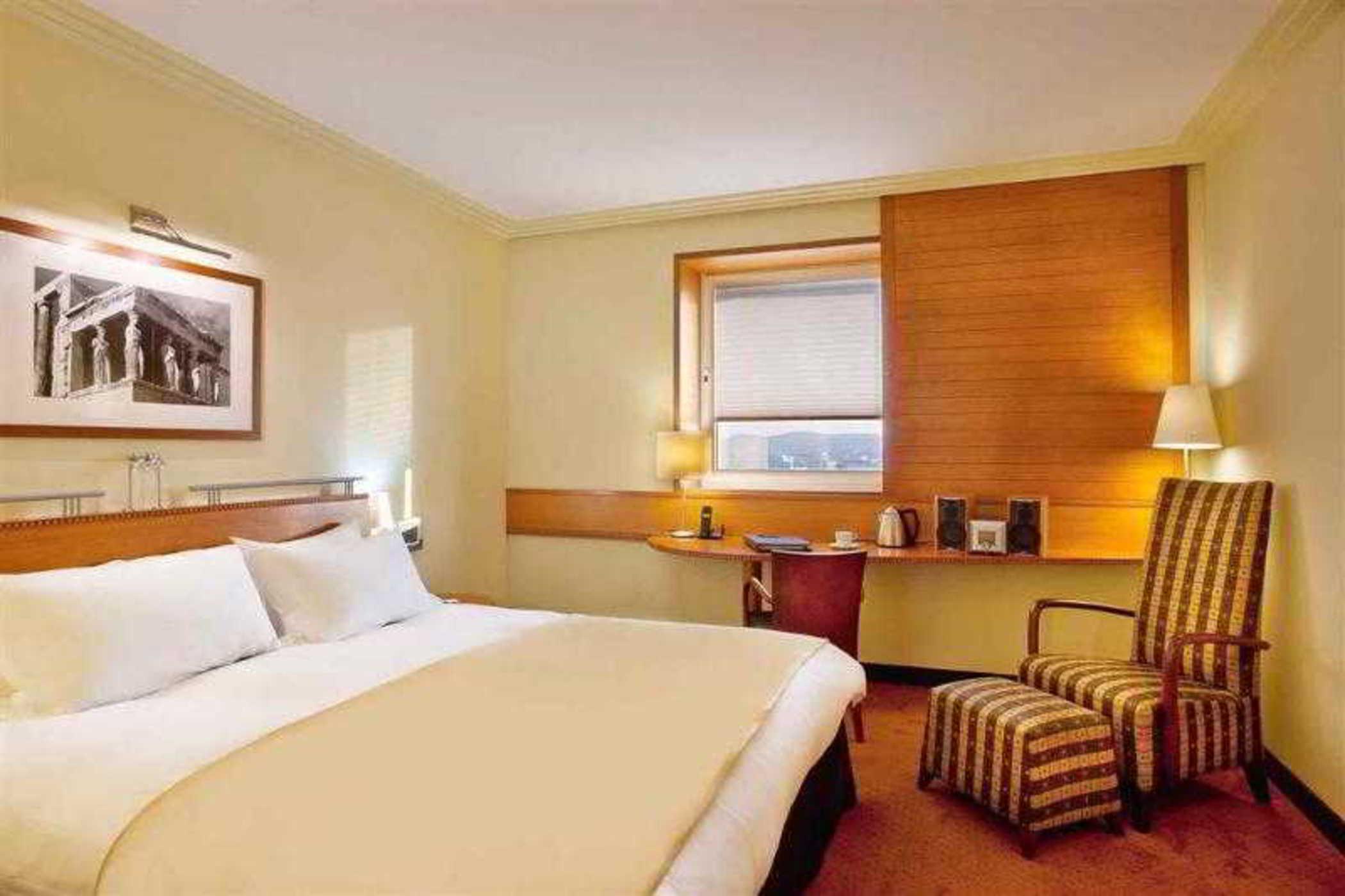 Sofitel Athens Airport Hotel Spata  Luaran gambar The photo depicts the interior of a hotel room. It features a large bed with white linens and a beige blanket. There is a window with blinds, allowing some natural light to enter the space. Beside the bed, there is a bedside lamp and a piece of artwo