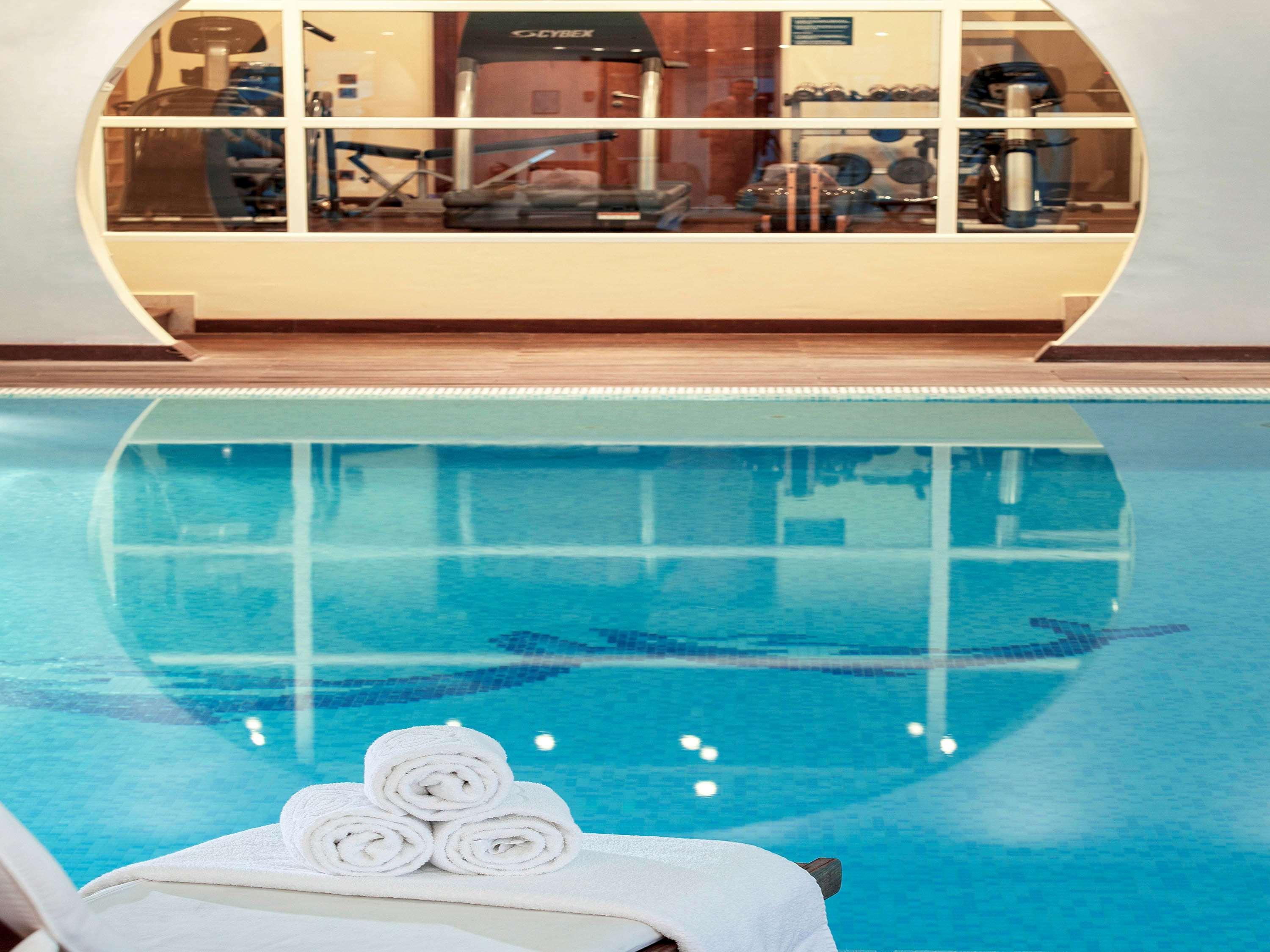 Sofitel Athens Airport Hotel Spata  Luaran gambar The photo shows a tranquil indoor swimming pool area. The water is clear and blue, and there are neatly stacked white towels on a lounge chair. In the background, there is a glimpse of a fitness area visible through large glass windows, featuring var