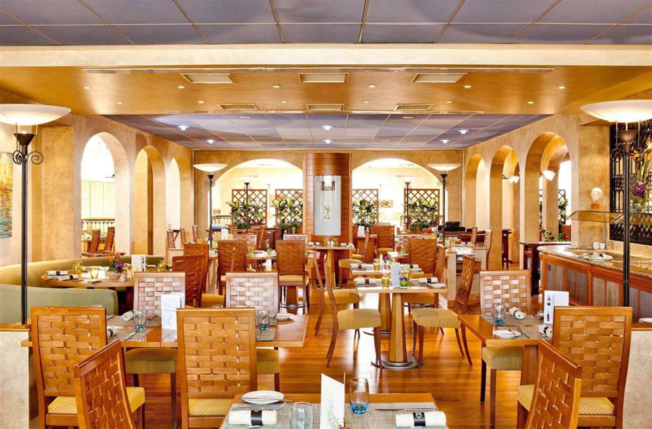 Sofitel Athens Airport Hotel Spata  Luaran gambar The photo shows the interior of a restaurant. The space features warm, golden tones with wooden furniture. There are several tables with chairs arranged throughout the room, and the tables appear to be set for dining with some plates and utensils. Ar