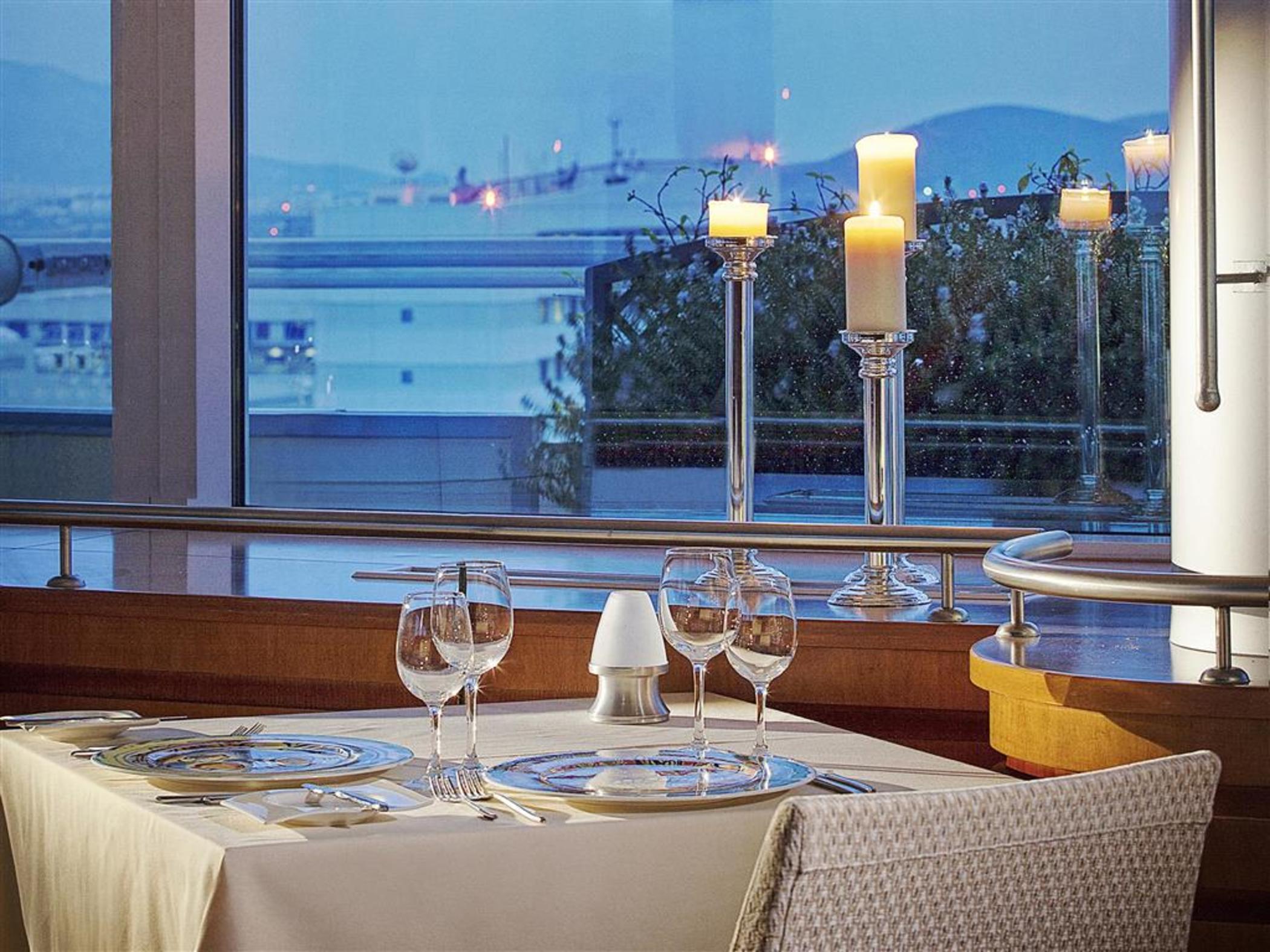 Sofitel Athens Airport Hotel Spata  Luaran gambar The photo shows an elegant dining setup in a restaurant. The table is covered with a light-colored tablecloth and is beautifully arranged with glassware, including wine glasses and a small salt shaker. In the background, there are large windows that 