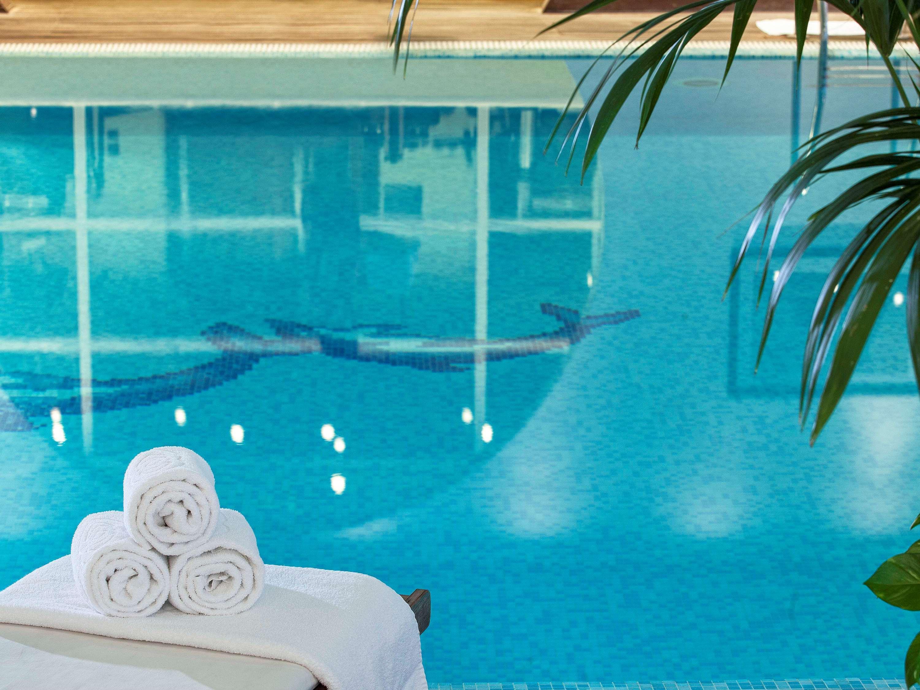 Sofitel Athens Airport Hotel Spata  Luaran gambar The photo shows a serene swimming pool with clear, turquoise water. In the foreground, there are neatly rolled towels placed on a lounge chair. The water reflects soft lighting and has a tiled pattern visible beneath the surface, while there are hint