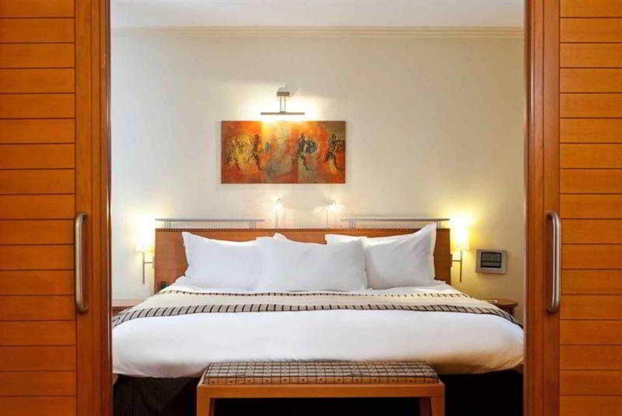 Sofitel Athens Airport Hotel Spata  Luaran gambar The photo shows a well-decorated hotel room with a contemporary design. At the center, there is a large bed with a white comforter and several pillows. Behind the bed, there is a piece of artwork featuring figures, which adds a pop of color to the ro