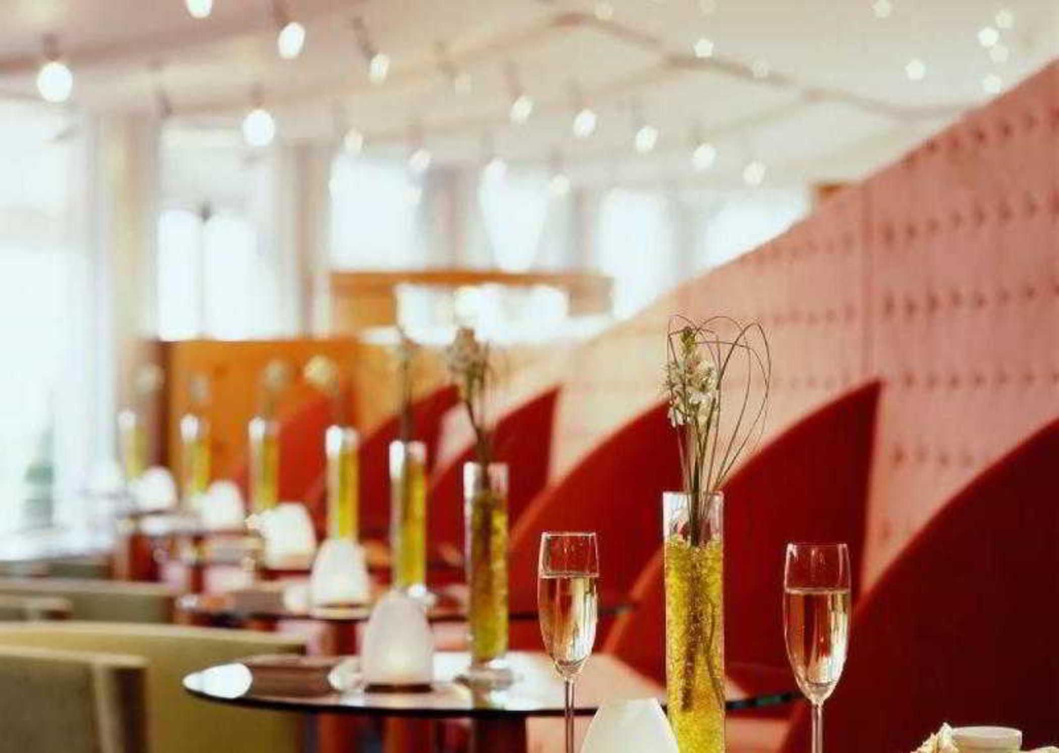 Sofitel Athens Airport Hotel Spata  Luaran gambar The image depicts the interior of a modern restaurant or lounge. The space features elegantly arranged tables with small vases of greenery as centerpieces. Each table has stemmed glasses, possibly for champagne or cocktails. The decor includes soft, 