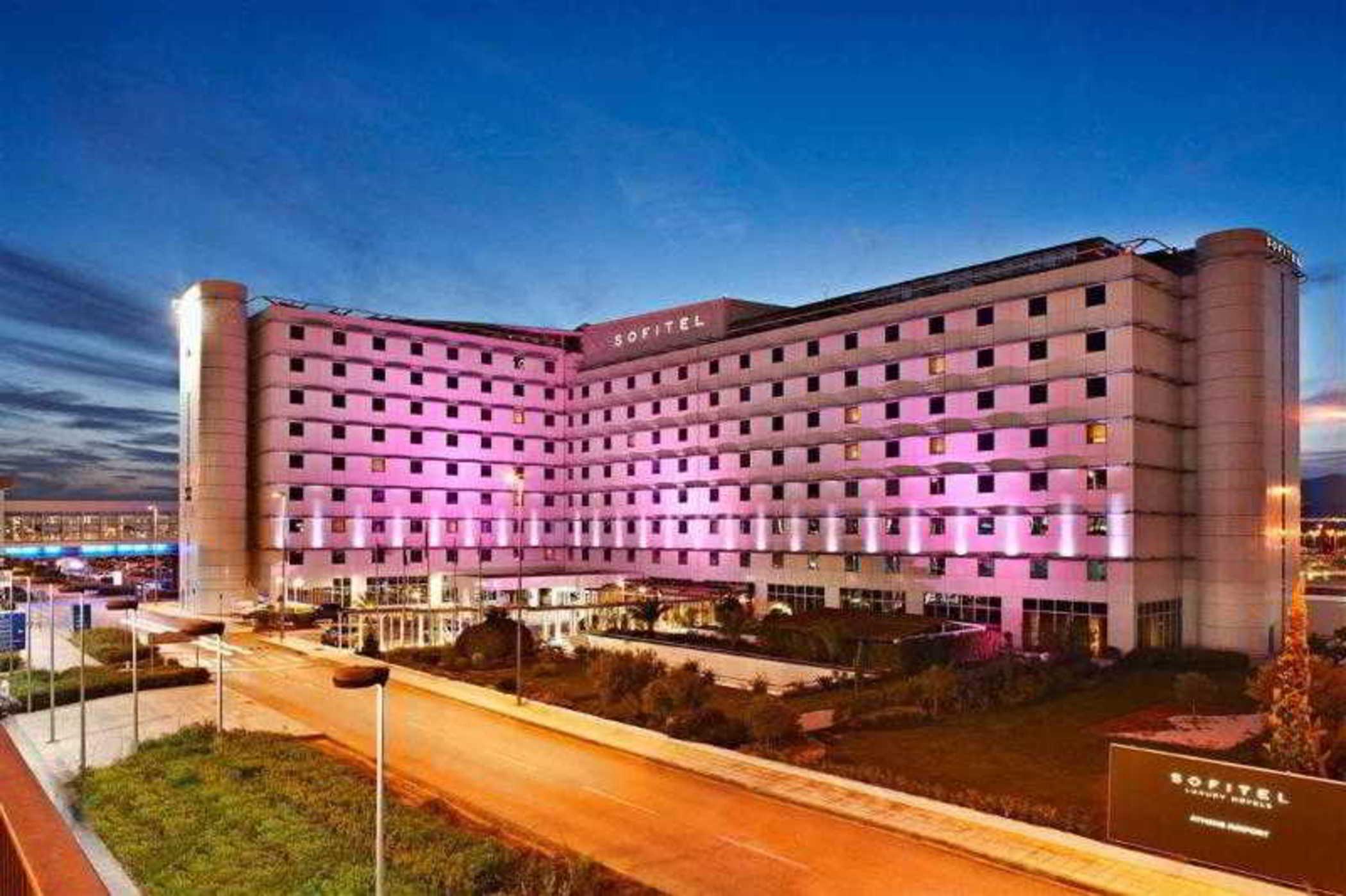 Sofitel Athens Airport Hotel Spata  Luaran gambar The photo displays a modern hotel building, likely a hotel brand, with a sleek, contemporary architectural design. The façade is illuminated in pink and purple lights, suggesting a vibrant atmosphere. The building has multiple floors, with large wind