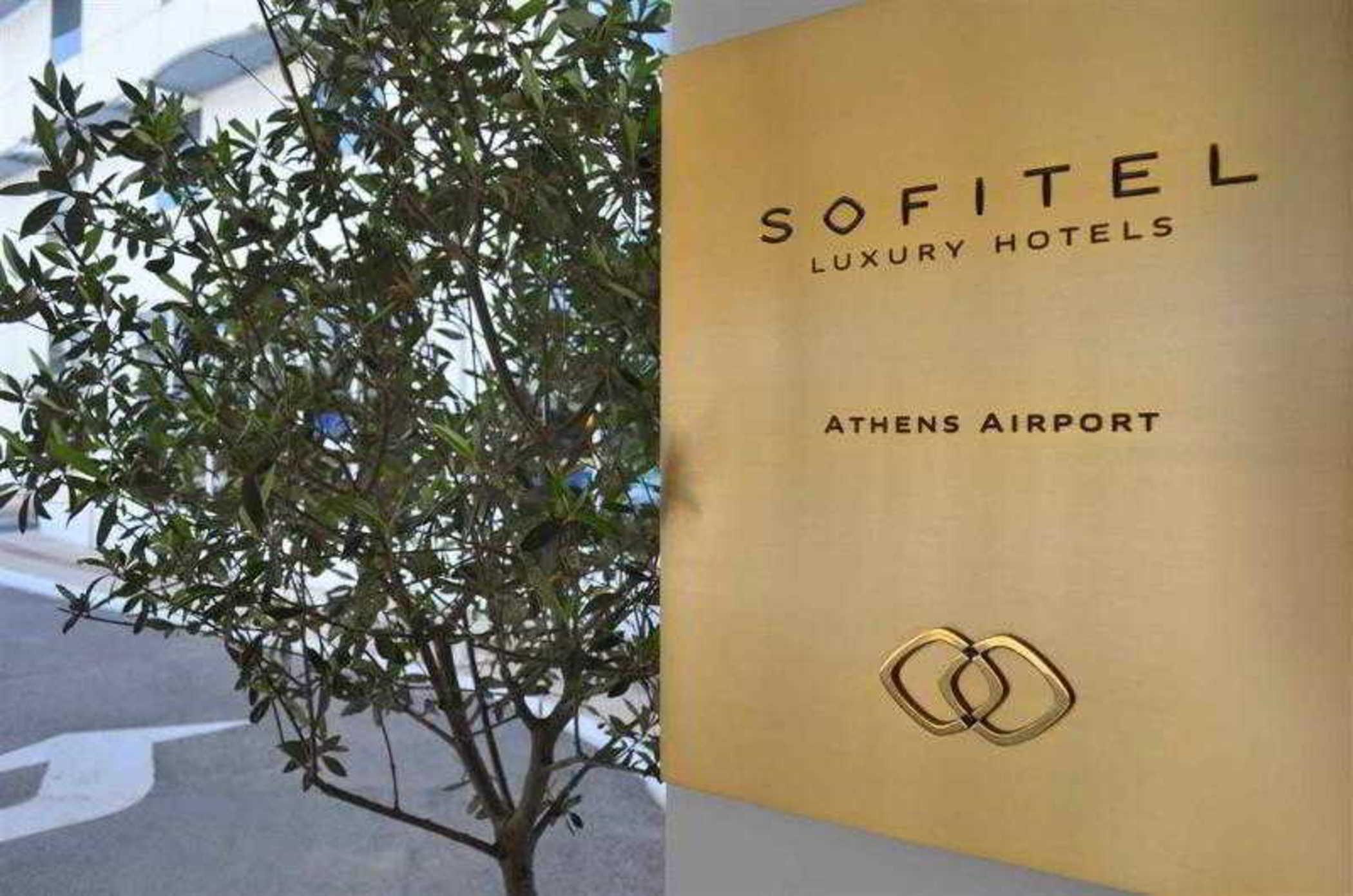 Sofitel Athens Airport Hotel Spata  Luaran gambar The photo shows two main elements. On the left side, there is a green tree with numerous leaves, which suggests a natural or outdoor setting. On the right side, there is a golden sign displaying the name "Sofitel Luxury Hotels" along with the text "A