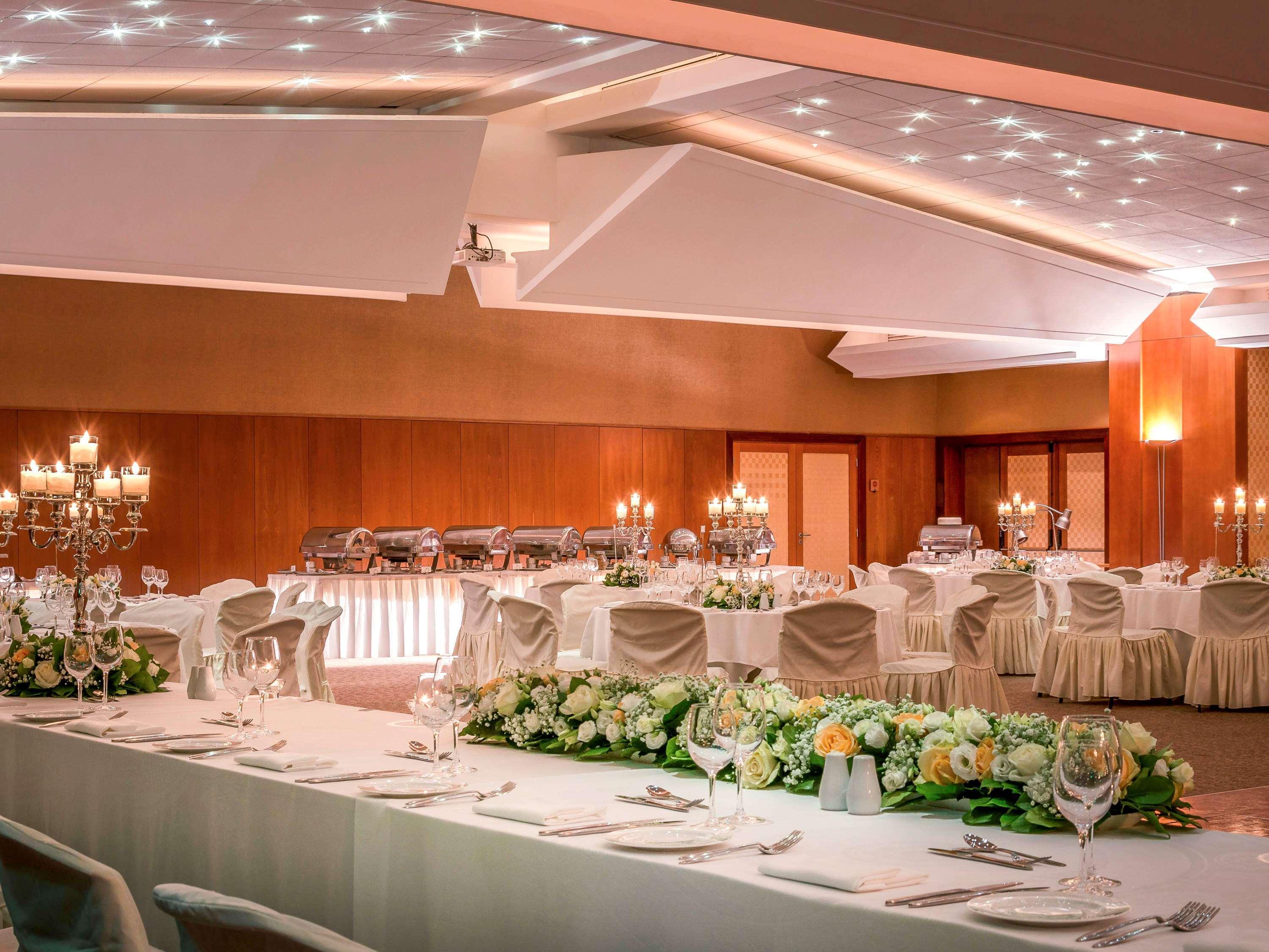 Sofitel Athens Airport Hotel Spata  Luaran gambar The photo shows an elegantly decorated banquet or event hall. The room features a large table set with fine dining ware, including crystal glasses and silver utensils. There are multiple round tables arranged around the space, each adorned with white
