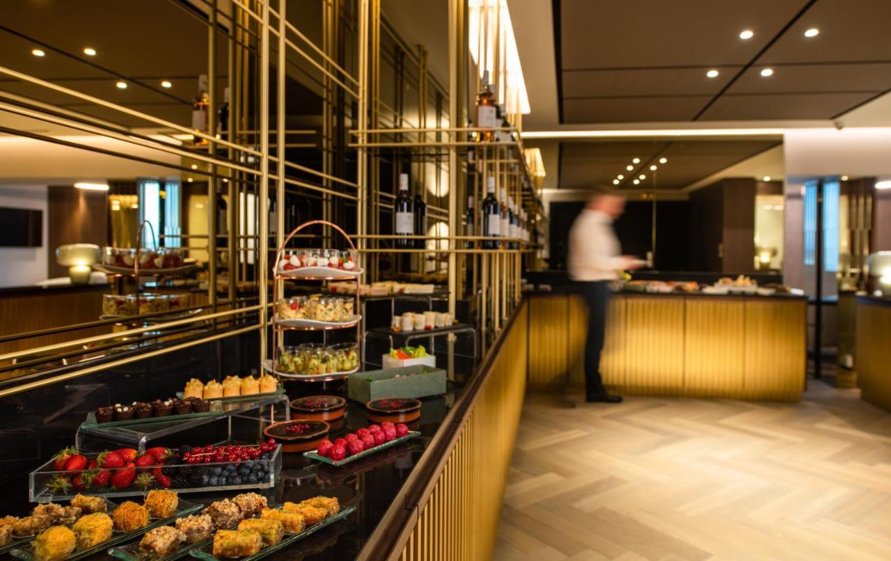Sofitel Athens Airport Hotel Spata  Luaran gambar The photo shows an elegantly designed buffet setup in a stylish interior. There are multiple displays of desserts and pastries on a black countertop, featuring items like fruits, cakes, and sweets arranged on glass trays. Behind the buffet, there are