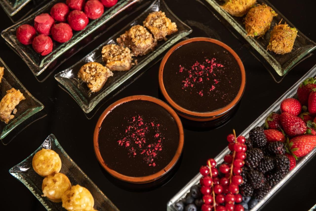 Sofitel Athens Airport Hotel Spata  Luaran gambar The image shows a beautifully arranged assortment of desserts and sweets on a dark surface. There are two brown bowls filled with a thick, dark dessert, garnished with rose petals. Surrounding the bowls are various plates containing a variety of trea