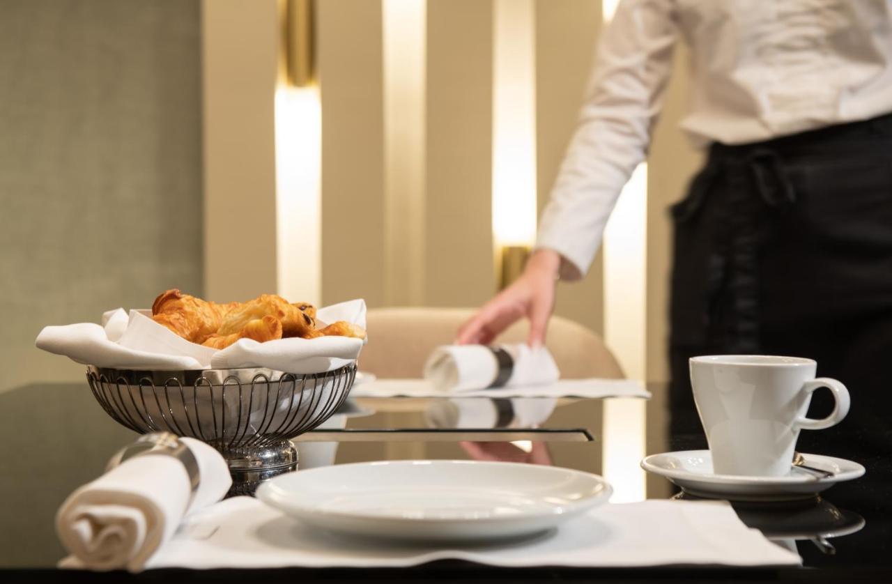 Sofitel Athens Airport Hotel Spata  Luaran gambar The photo shows a dining table setting in a restaurant. On the table, there is a white plate, a small cup, and a silver bowl containing pastries, likely croissants. The surrounding area has a clean, modern aesthetic with soft lighting. In the backgro