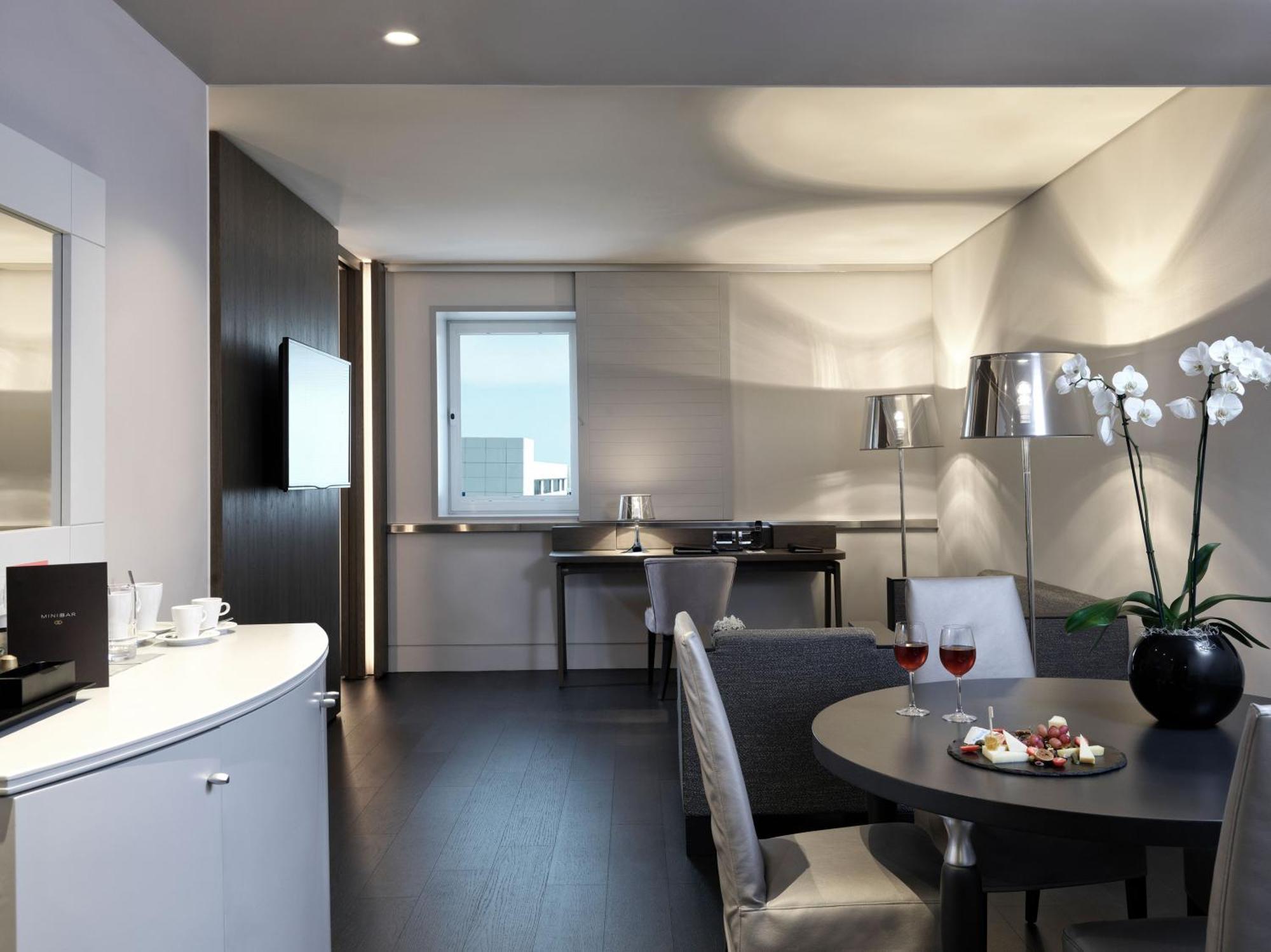 Sofitel Athens Airport Hotel Spata  Luaran gambar The photo shows a modern and elegantly furnished living space, likely a hotel room or a suite. The room features:

- A stylish seating area with a gray armchair and a small table.
- A round dining table set for two, with plates of food and drinks.
- 
