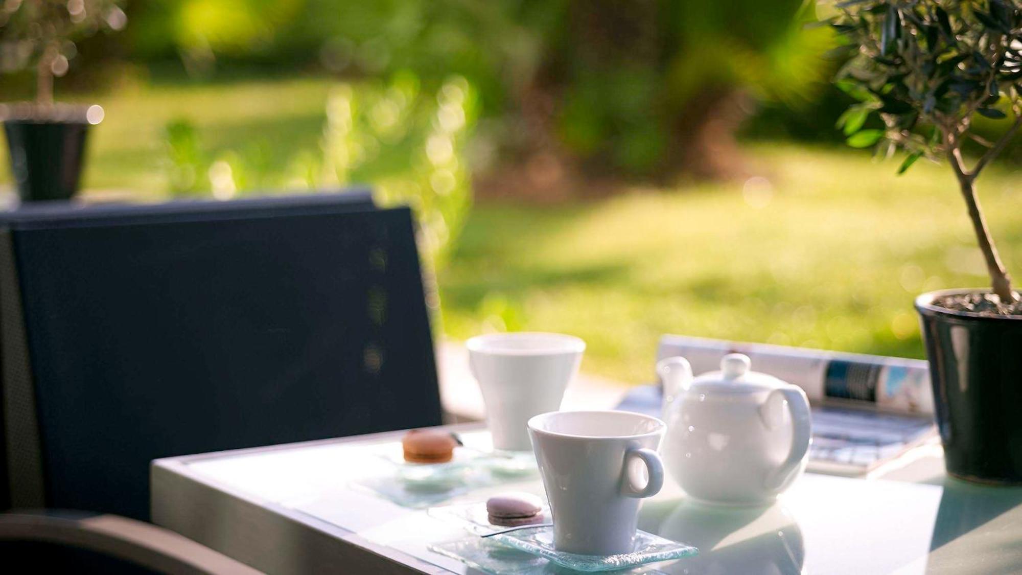 Sofitel Athens Airport Hotel Spata  Luaran gambar The photo shows a serene outdoor setting with a table featuring a tea setup. There are two white cups, a white teapot, and a small cookie or biscuit placed on the table. The background features lush greenery, suggesting a tranquil garden or patio atm