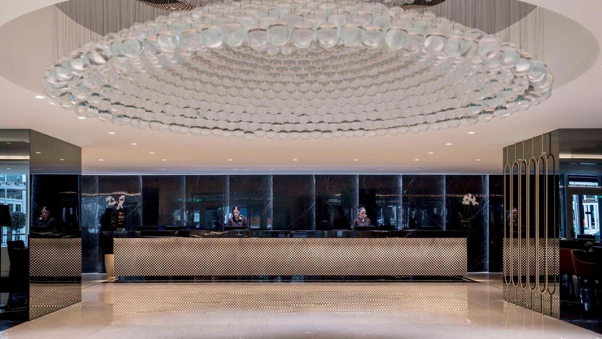Sofitel Athens Airport Hotel Spata  Luaran gambar The photo shows a modern hotel lobby. The space features a sleek design with a large, artistic chandelier made of glass or crystal that hangs from the ceiling. The reception desk is centrally located and designed with a polished, reflective surface. 