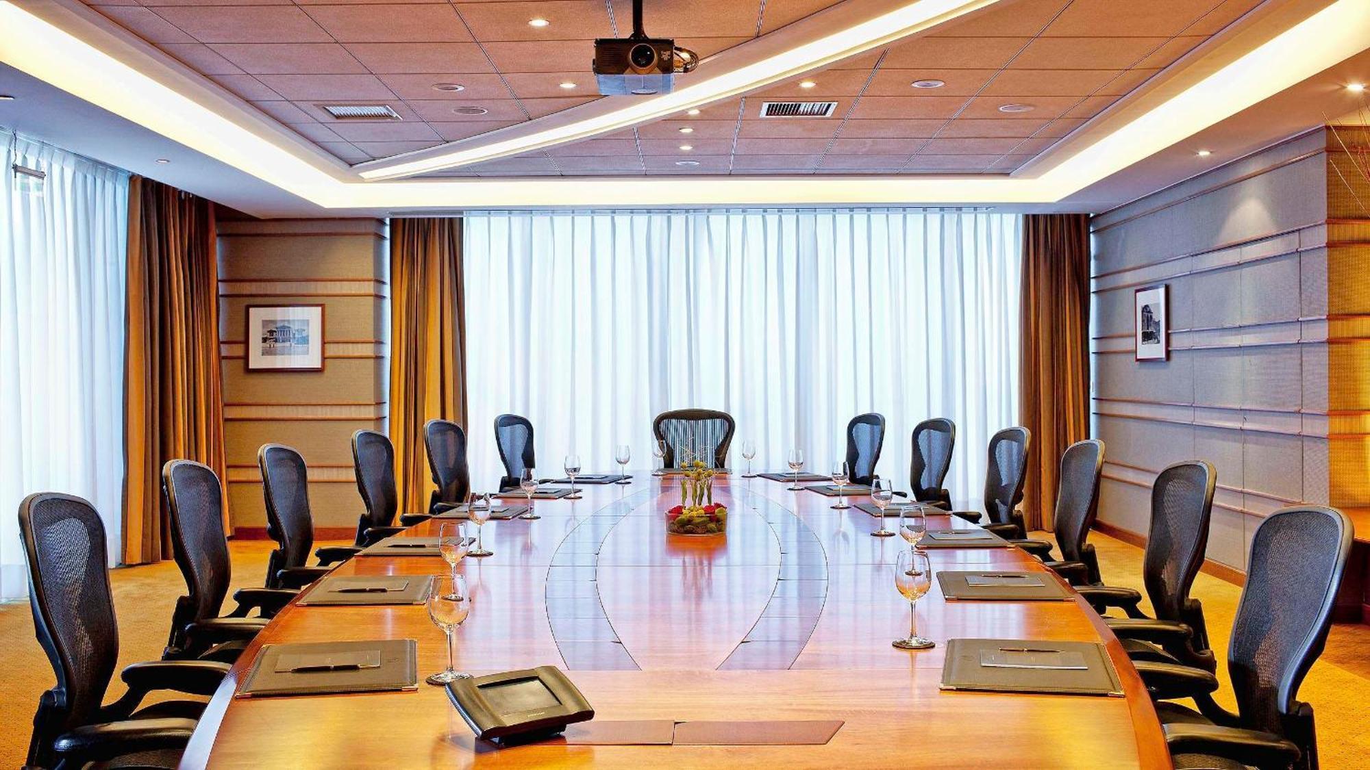Sofitel Athens Airport Hotel Spata  Luaran gambar The photo depicts a modern conference room featuring a large, oval wooden table surrounded by several ergonomic chairs. The room has dim lighting and is illuminated by natural light coming in through large windows covered by sheer curtains. There are