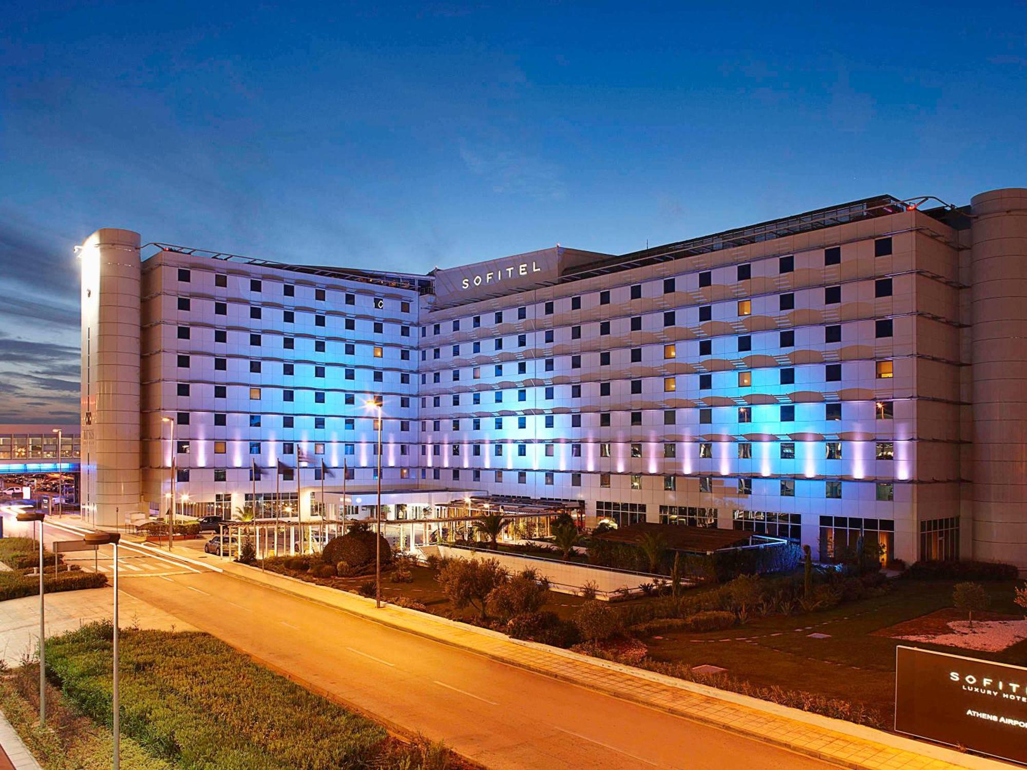 Sofitel Athens Airport Hotel Spata  Luaran gambar The photo shows a large modern hotel building, likely named "Sofitel," illuminated with blue and purple lights as evening approaches. The structure features multiple floors and a contemporary design, with large windows and a flat facade. In front of 
