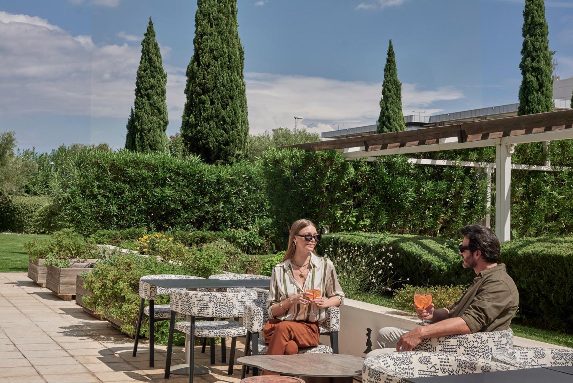 Sofitel Athens Airport Hotel Spata  Luaran gambar The photo shows two people seated at an outdoor setting, enjoying drinks. They are positioned at a table with decorative tablecloths, surrounded by lush greenery and tall cypress trees. The atmosphere appears relaxed and sunny, suggesting a pleasant 