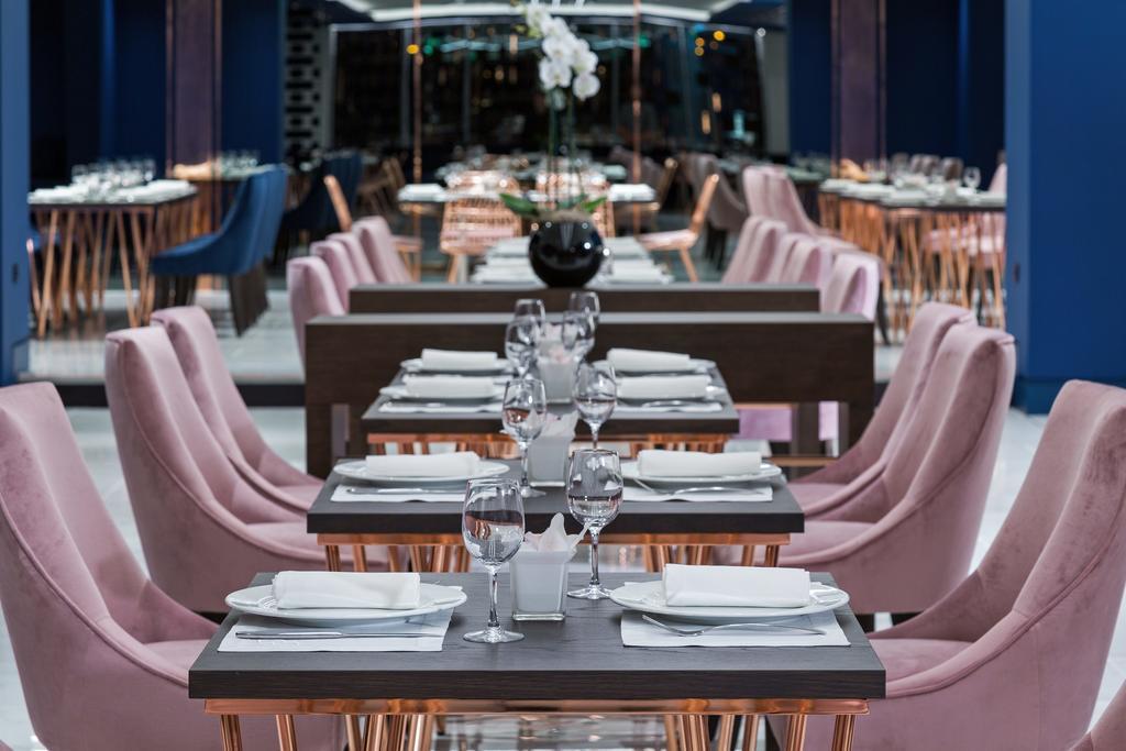 Sofitel Athens Airport Hotel Spata  Luaran gambar The photo showcases a modern restaurant interior. In the foreground, there are several tables set with white plates, glassware, and napkins, arranged neatly. Each table has elegant pink chairs, giving a chic and contemporary vibe. In the background, 