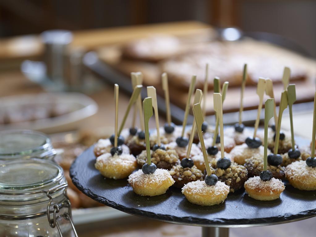 Sofitel Athens Airport Hotel Spata  Luaran gambar The photo displays a dessert selection featuring small pastries arranged on a slate serving platter. The pastries are topped with coconut and have a single blue berry on each, with decorative sticks inserted for easy handling. In the background, ther