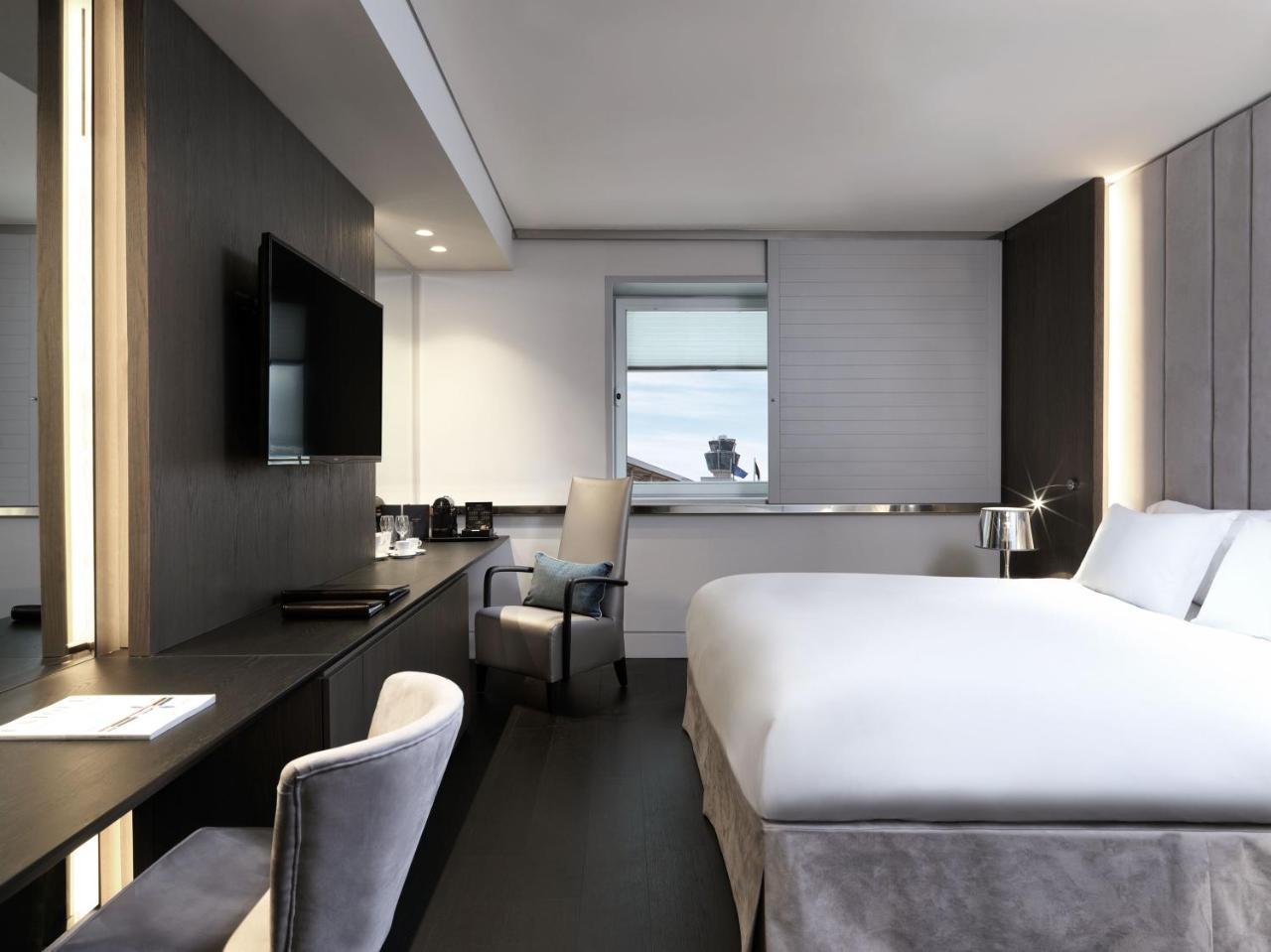 Sofitel Athens Airport Hotel Spata  Luaran gambar The photo shows a modern hotel room. It features a large bed with white bedding, a sleek desk with a chair, and a flat-screen TV mounted on the wall. There are large windows allowing natural light to enter, and the room is decorated in neutral tones 